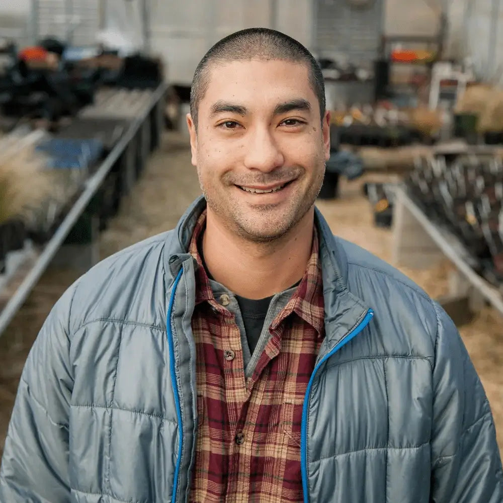 Man wearing a gray puffer jacket and plaid shirt in an indoor setting with plants and equipment.